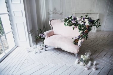 Çiçekler ve yeşillerle süslenmiş, yumuşak pembe renkli kanepe, büyük pencere ve perdelerin yanındaki cam şamdanlarla çevrili beyaz ahşap bir zeminde klasik bir odada duruyor.