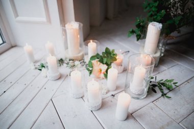 Cam şamdanların içindeki beyaz mumların güzel dekoru yeşil yaprakların ve çiçeklerin arasında beyaz ahşap bir zeminde duruyor.