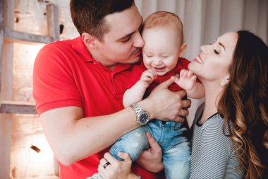 Anne ve baba, ellerinde aynı kotlu ve kırmızı tişörtlü küçük bir çocuk, parlak bir odada, perdeleri, merdivenleri ve ampulleri olan beyaz tuğladan bir duvara karşı duruyor.