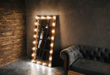 Köşe odasında ahşap ve gri beton bir duvar, yaşlanmış bir zemin, siyah cam kapılar ve ampullü elektrikli makyaj aynası ve tavan arası tarzı Oxford kanepesi.