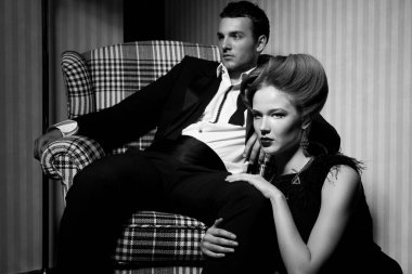 Odadaki gölgelerde klasik giysiler içinde tutkulu güzel bir çift: sandalyede oturan adam, ayaklarının dibinde yatan kadın.