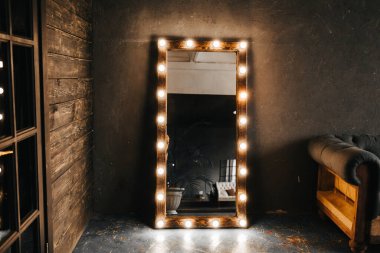 Köşe odasında ahşap ve gri beton bir duvar, yaşlanmış bir zemin, siyah cam kapılar ve ampullü elektrikli makyaj aynası ve tavan arası tarzı Oxford kanepesi.