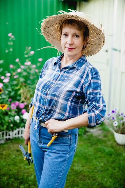 stylish aged woman retired gardener in blue jeans, checkered shirt and straw hat stay on background of a wooden gazebo