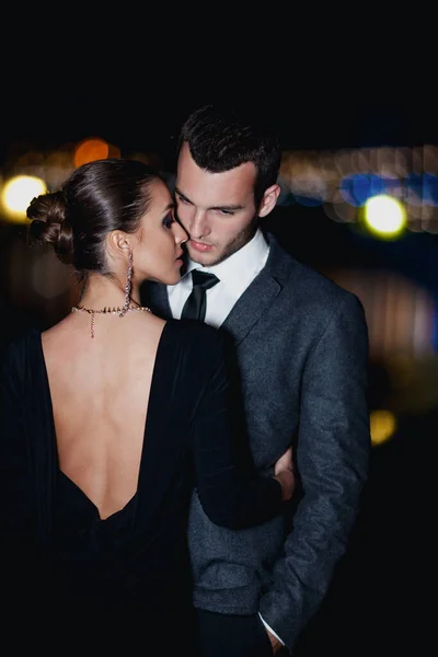 Şık giyinmiş zarif bir çift akşam vakti dışarıda sarılıyor.