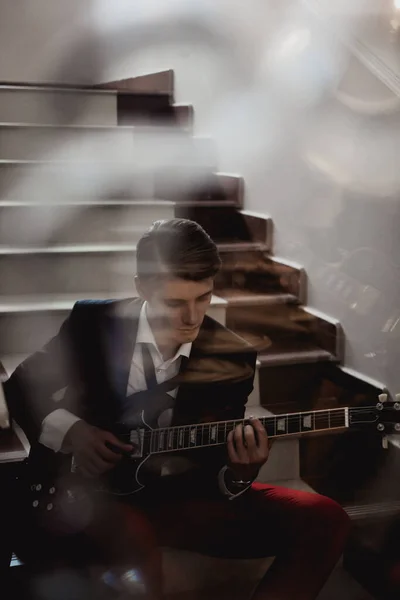 階段に座って鏡の反射でギターを弾く赤いズボンと黒いジャケットのハンサムな男 — ストック写真