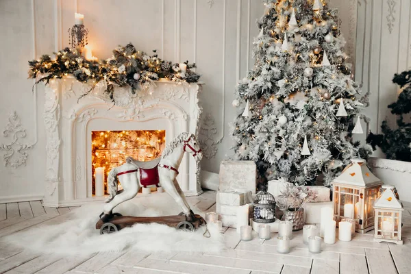 Beyaz bir odanın içinde Noel ağacı, süslü bir şömine, sallanan at ve yeni yıl hediyeleri olan bir oda.