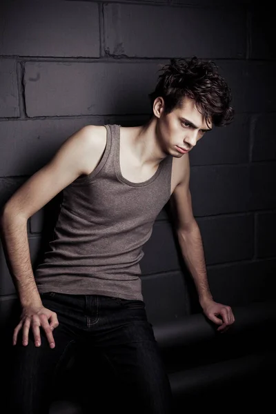 A young thin dark-haired guy in a gray T-shirt posing against a concrete-brick wall