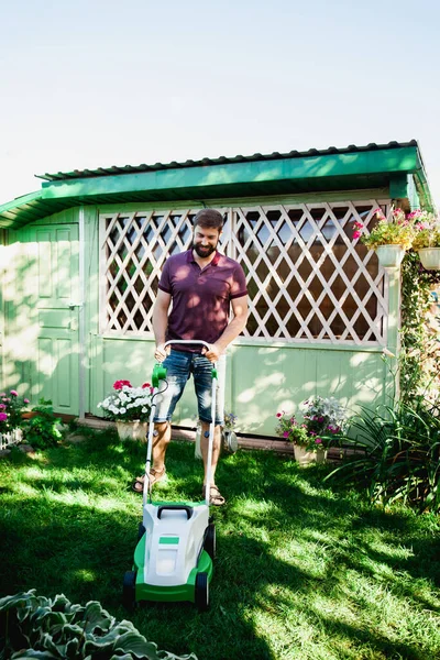 tall handsome happy bearded man in jeans and polo pushes forward lawnmower on green grass in the backyard of a country house and smiles on a sunny summer day against the backdrop of a wooden gazebo