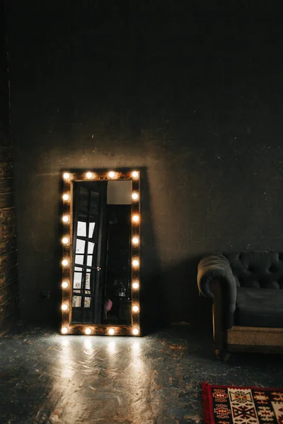 Corner room with a wooden and gray cement wall, an aged floor, black glass doors and electric dressing makeup mirror with lights bulbs and oxford sofa in loft style