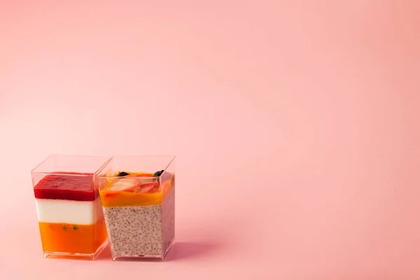 two juicy fresh desserts in cups pudding, chia seeds, mango puree, berries, multi-layer, panakota, passion fruit coconut milk on a pink background in isolated. copy space, place for text