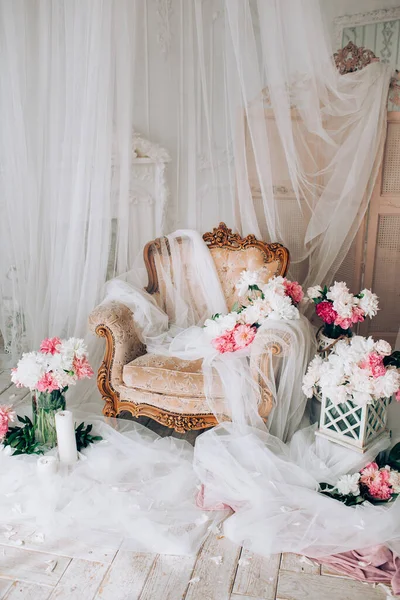 vintage armchair decorated with peonies flowers and greens, stands in a classic room on a white wooden floor surrounded by candles near large window and curtains