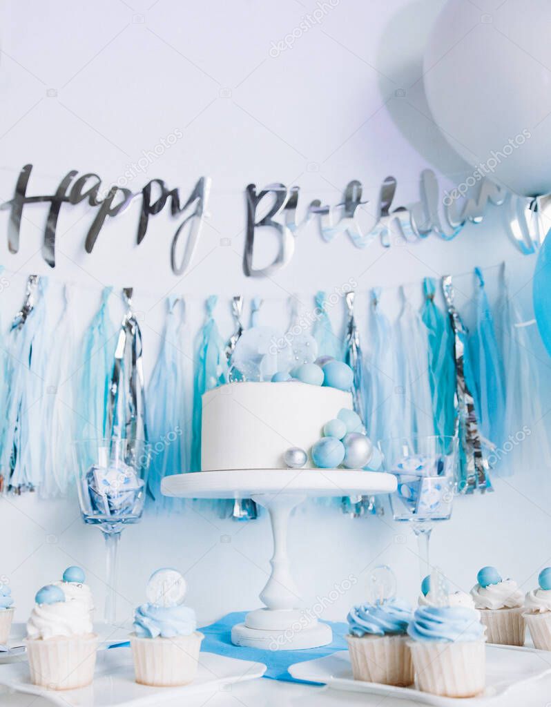 festive decor in honor of one year old baby boy. white cake on a stand, cupcakes, blue balls and tinsel. the inscription 
