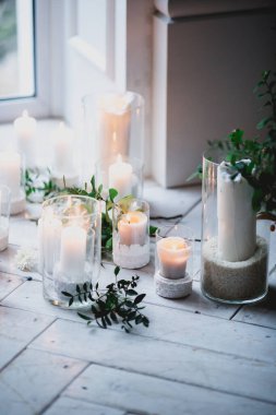 Cam şamdanların içindeki beyaz mumların güzel dekoru yeşil yaprakların ve çiçeklerin arasında beyaz ahşap bir zeminde duruyor.