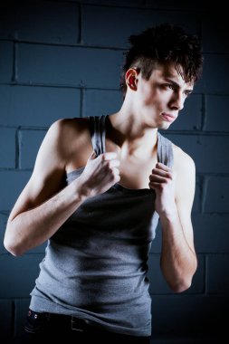 Gri tişörtlü, esmer, genç bir adam beton duvara karşı poz veriyor.