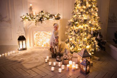 Beyaz elbiseli sevimli çocuk Noel ağacı ve şömineli at oyuncağının üstünde.