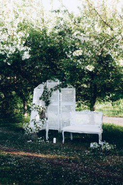 Yeşil bir parkta güzel bir düğün fotoğrafı. Gölgede elma ağaçları, beyaz bank, ekran, çiçekli kaide ve dekor.