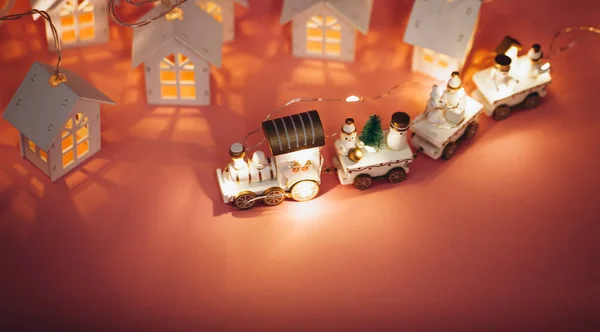 Holiday magic concept of New Year small toy miniature white train with Santa, snowman, garland lights, Christmas tree,small houses with luminous windows on pink background