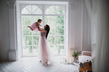 Güzel esmer kadın. Elbise giymiş anne sevgili kızıyla çiçeklerle ve mumlarla süslenmiş parlak beyaz bir odada oynuyor.