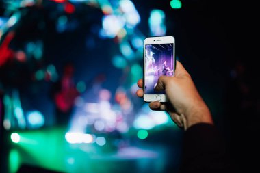 Konserdeki hayran elinde bir akıllı telefon tutuyor ve fotoğraf çekiyor ya da arka planda video çekiyor. Bir şarkıcı ve müzik grubunun bulanık performansı.