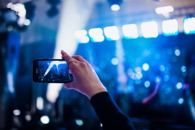 Konserdeki hayran elinde bir akıllı telefon tutuyor ve fotoğraf çekiyor ya da arka planda video çekiyor. Bir şarkıcı ve müzik grubunun bulanık performansı.
