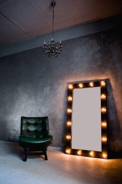 yeşil deri koltuk ve uzun giyinme aynası. Koyu renkli bir odada, gri bir duvarın arka planında, üzerinde lambalar olan bir çerçeve.
