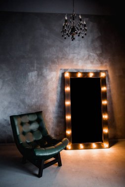 yeşil deri koltuk ve uzun giyinme aynası. Koyu renkli bir odada, gri bir duvarın arka planında, üzerinde lambalar olan bir çerçeve.