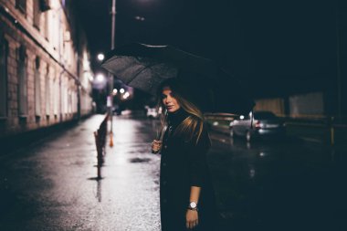 Güzel, genç, yalnız, sarışın bir kız. Gece caddesinde şemsiyenin altında duruyor.