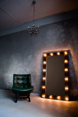 yeşil deri koltuk ve uzun giyinme aynası. Koyu renkli bir odada, gri bir duvarın arka planında, üzerinde lambalar olan bir çerçeve.