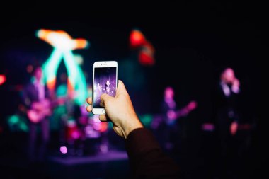 Konserdeki hayran elinde bir akıllı telefon tutuyor ve fotoğraf çekiyor ya da arka planda video çekiyor. Bir şarkıcı ve müzik grubunun bulanık performansı.
