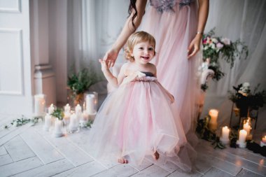 Anne küçük bir kız çocuğu tutuyor, ilk adımlarını atıyor, pembe güzel elbiseler içinde, çiçeklerle ve mumlarla süslenmiş beyaz bir stüdyo odasında, mutlu güzel bir fotoğraf.