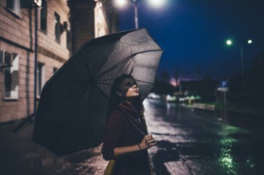 Güzel, genç, yalnız, sarışın bir kız. Gece caddesinde şemsiyenin altında duruyor.