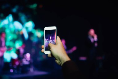 Konserdeki hayran elinde bir akıllı telefon tutuyor ve fotoğraf çekiyor ya da arka planda video çekiyor. Bir şarkıcı ve müzik grubunun bulanık performansı.