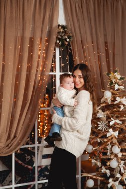 Mutlu anne, oğlunun kollarında rahat kazaklar içinde şeffaf bir perdesi, ışıkları ve Noel ağacı olan bir pencerenin arkasında duruyor.