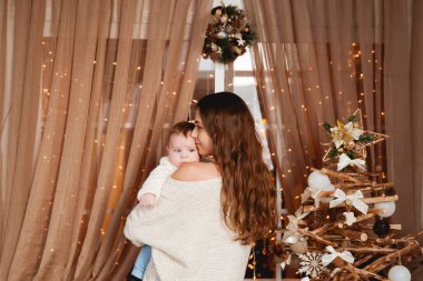 Mutlu anne, oğlunun kollarında rahat kazaklar içinde şeffaf bir perdesi, ışıkları ve Noel ağacı olan bir pencerenin arkasında duruyor.