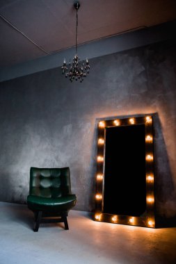yeşil deri koltuk ve uzun giyinme aynası. Koyu renkli bir odada, gri bir duvarın arka planında, üzerinde lambalar olan bir çerçeve.