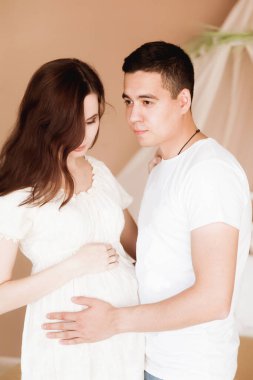 Gelecekteki mutlu ebeveynler beyaz bir yatağın arka planında dururlar.