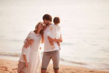 Beyaz elbiseli mutlu aile gün batımında sahilde duruyor.