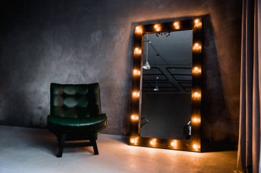 yeşil deri koltuk ve uzun giyinme aynası. Koyu renkli bir odada, gri bir duvarın arka planında, üzerinde lambalar olan bir çerçeve.