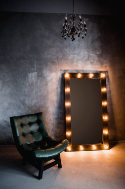 yeşil deri koltuk ve uzun giyinme aynası. Koyu renkli bir odada, gri bir duvarın arka planında, üzerinde lambalar olan bir çerçeve.