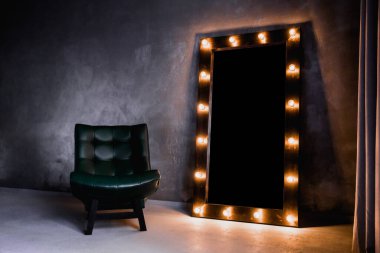 yeşil deri koltuk ve uzun giyinme aynası. Koyu renkli bir odada, gri bir duvarın arka planında, üzerinde lambalar olan bir çerçeve.