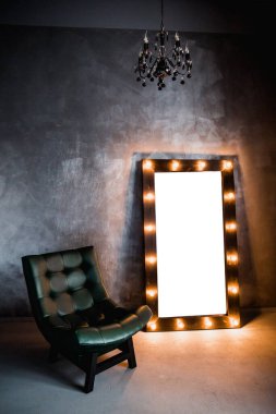 yeşil deri koltuk ve uzun giyinme aynası. Koyu renkli bir odada, gri bir duvarın arka planında, üzerinde lambalar olan bir çerçeve.