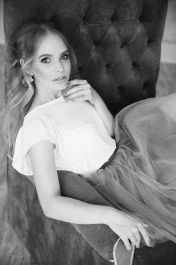 Güzel, sarışın bir manken. Gözleri pembe, gür bir etek ve beyaz bir bluz içinde koltukta oturuyor.