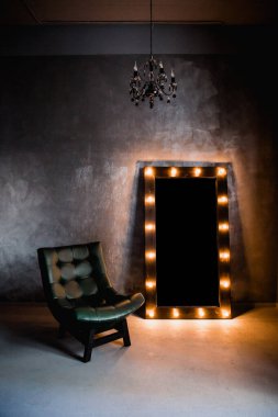 yeşil deri koltuk ve uzun giyinme aynası. Koyu renkli bir odada, gri bir duvarın arka planında, üzerinde lambalar olan bir çerçeve.