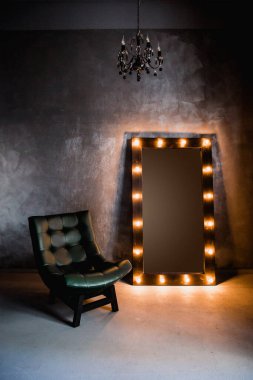 yeşil deri koltuk ve uzun giyinme aynası. Koyu renkli bir odada, gri bir duvarın arka planında, üzerinde lambalar olan bir çerçeve.