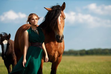Uzun yeşil elbiseli güzel sarışın kadın kahverengi at aygırları sürüsünün arasında bir tarlada duruyor. Güzel sanatsal duygusal fotoğraf, rüzgar elbisesini geliştiriyor, bulutlu gökyüzü.