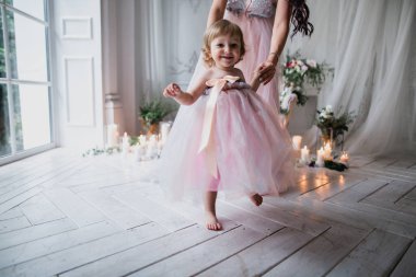 Anne küçük bir kız çocuğu tutuyor, ilk adımlarını atıyor, pembe güzel elbiseler içinde, çiçeklerle ve mumlarla süslenmiş beyaz bir stüdyo odasında, mutlu güzel bir fotoğraf.