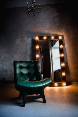 yeşil deri koltuk ve uzun giyinme aynası. Koyu renkli bir odada, gri bir duvarın arka planında, üzerinde lambalar olan bir çerçeve.