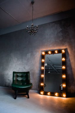 yeşil deri koltuk ve uzun giyinme aynası. Koyu renkli bir odada, gri bir duvarın arka planında, üzerinde lambalar olan bir çerçeve.