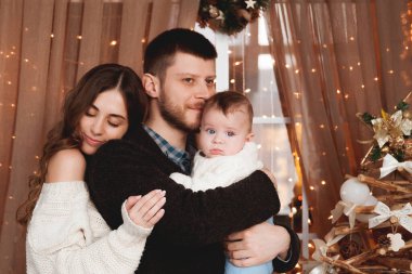 Oğulları kollarında rahat kazaklarla mutlu bir aile şeffaf bir perdesi, ışıkları ve Noel ağacı olan bir pencerenin arkasında dururlar.