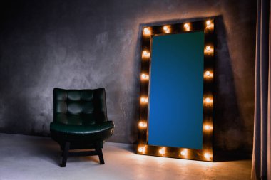 yeşil deri koltuk ve uzun giyinme aynası. Koyu renkli bir odada, gri bir duvarın arka planında, üzerinde lambalar olan bir çerçeve.